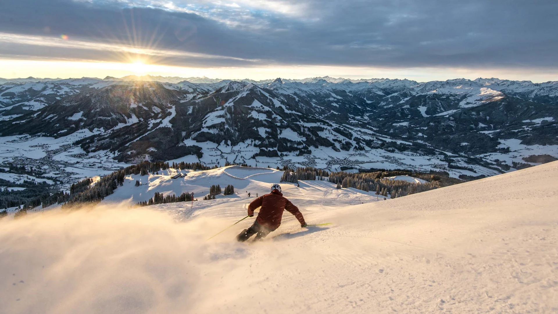 Skier skiing down snowy mountain at sunset with mountain range background