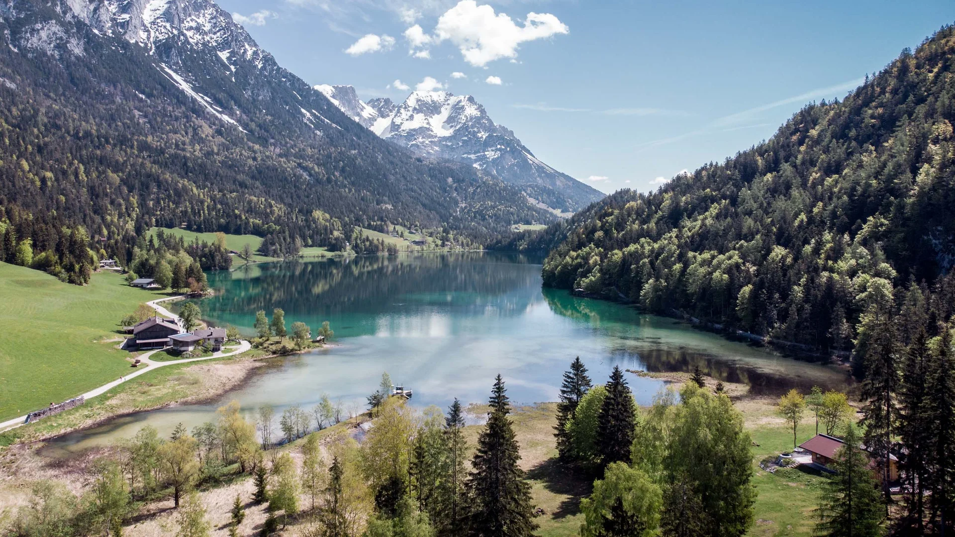 Bergsee mit schneebedeckten Gipfeln, umgeben von Wäldern und grünen Wiesen
