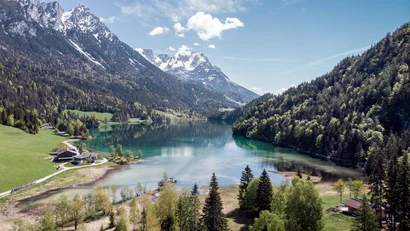 Bergsee mit schneebedeckten Gipfeln, umgeben von Wäldern und grünen Wiesen
